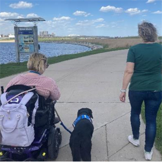 Two people walking along the lakefront, one person using a wheelchair with a service dog