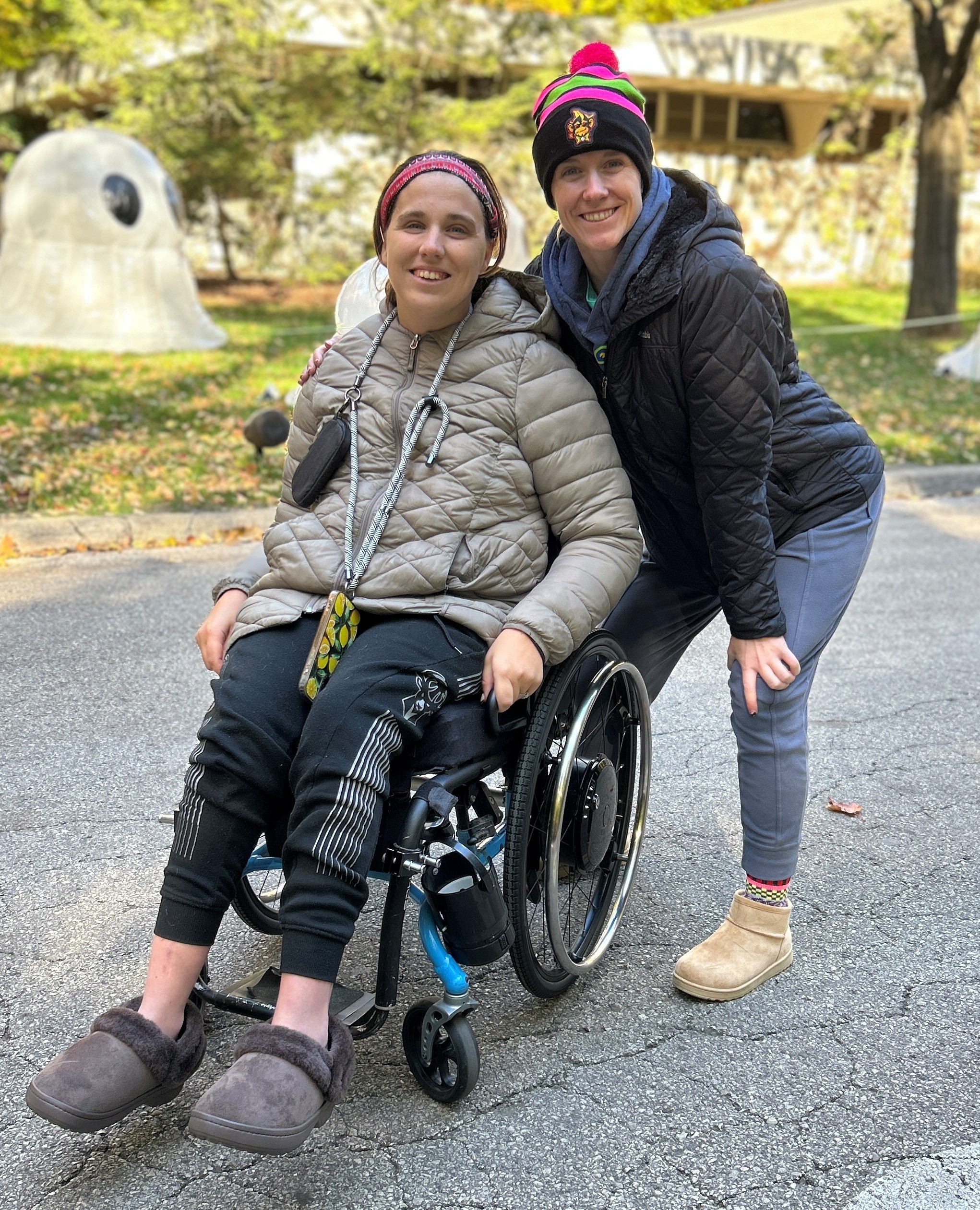 A young woman seated in a wheelchair smiles and poses for a photo. Another woman in a black coat bends down behind her also smiling.