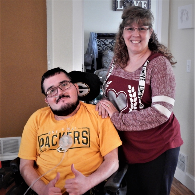 A man wearing a Packer shirt is sitting in a wheelchair and a woman wearing a red shirt with hearts is standing to the right of the wheelchair.  chair