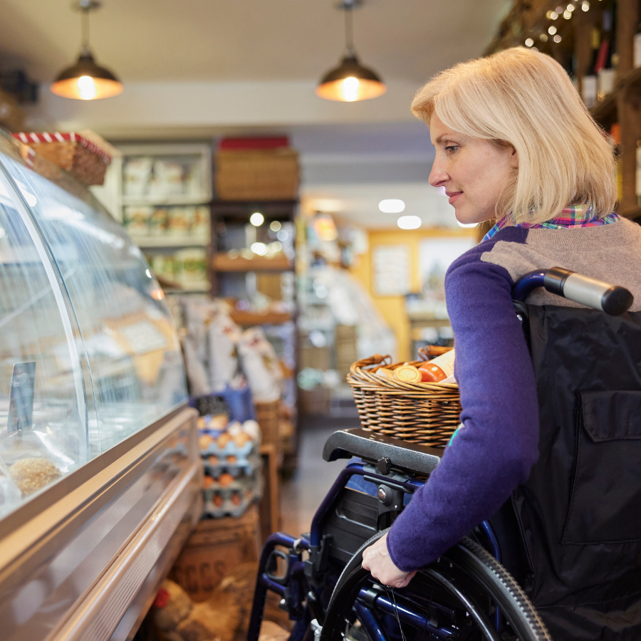 A woman looks into a deli case while seated in a wheelchair with a shopping basket in her lap