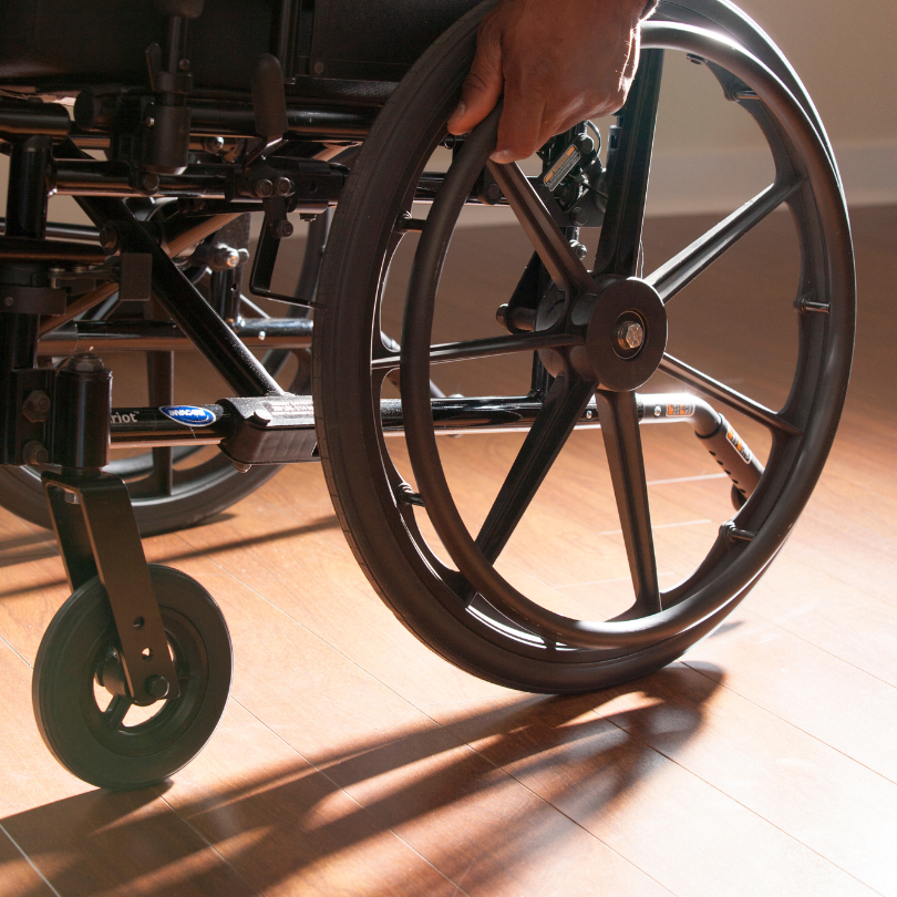 A close-up view of a wheelchair. A hand is gripping the larger wheel from above.