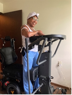 Dalphina wears a white tank top and blue pants with a white turban and glasses. She is smiling and standing with an assistive device