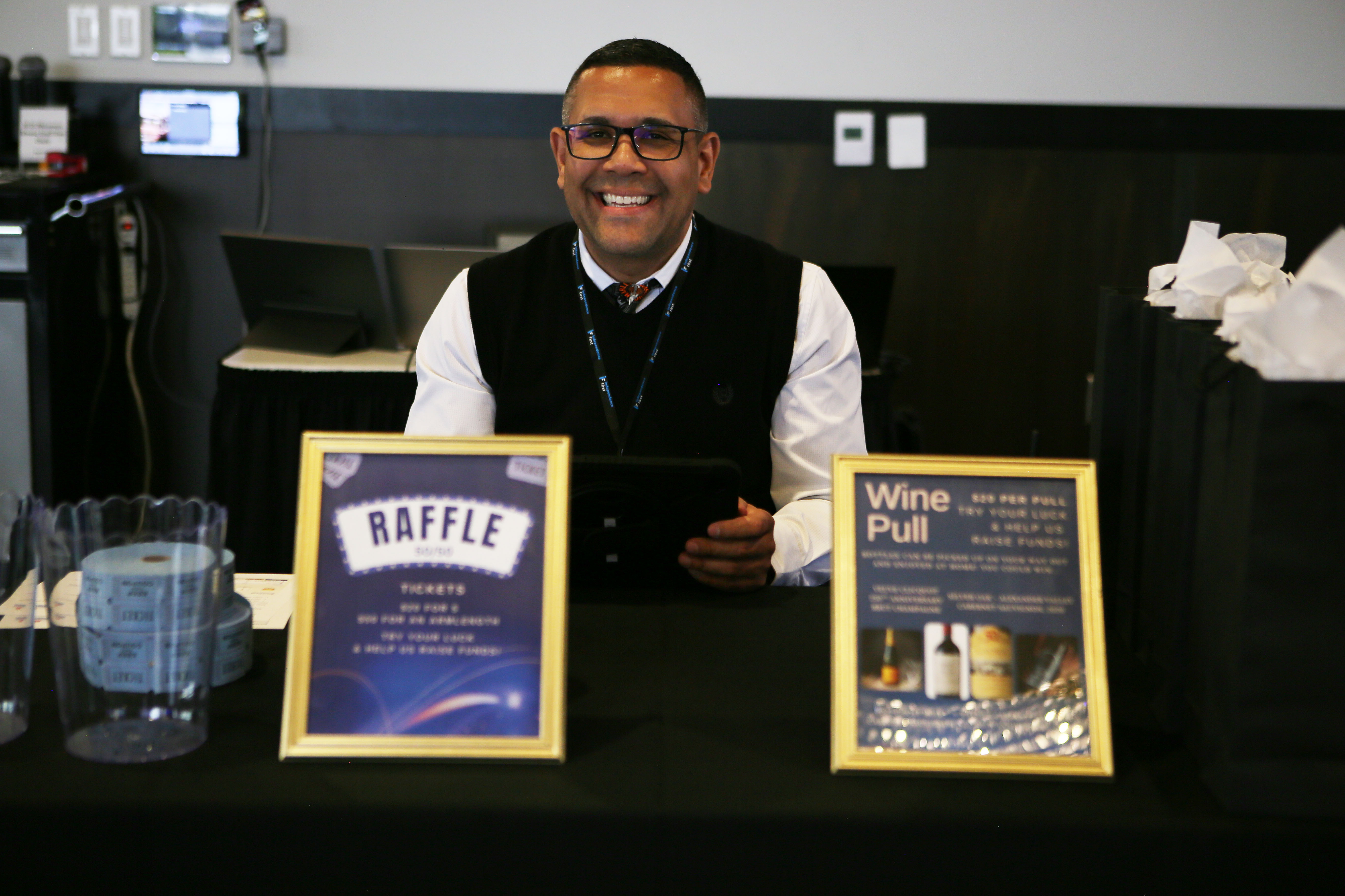 Independence First employee Eric Edwards smiles behind a table with 50/50 raffle and wine pull signs