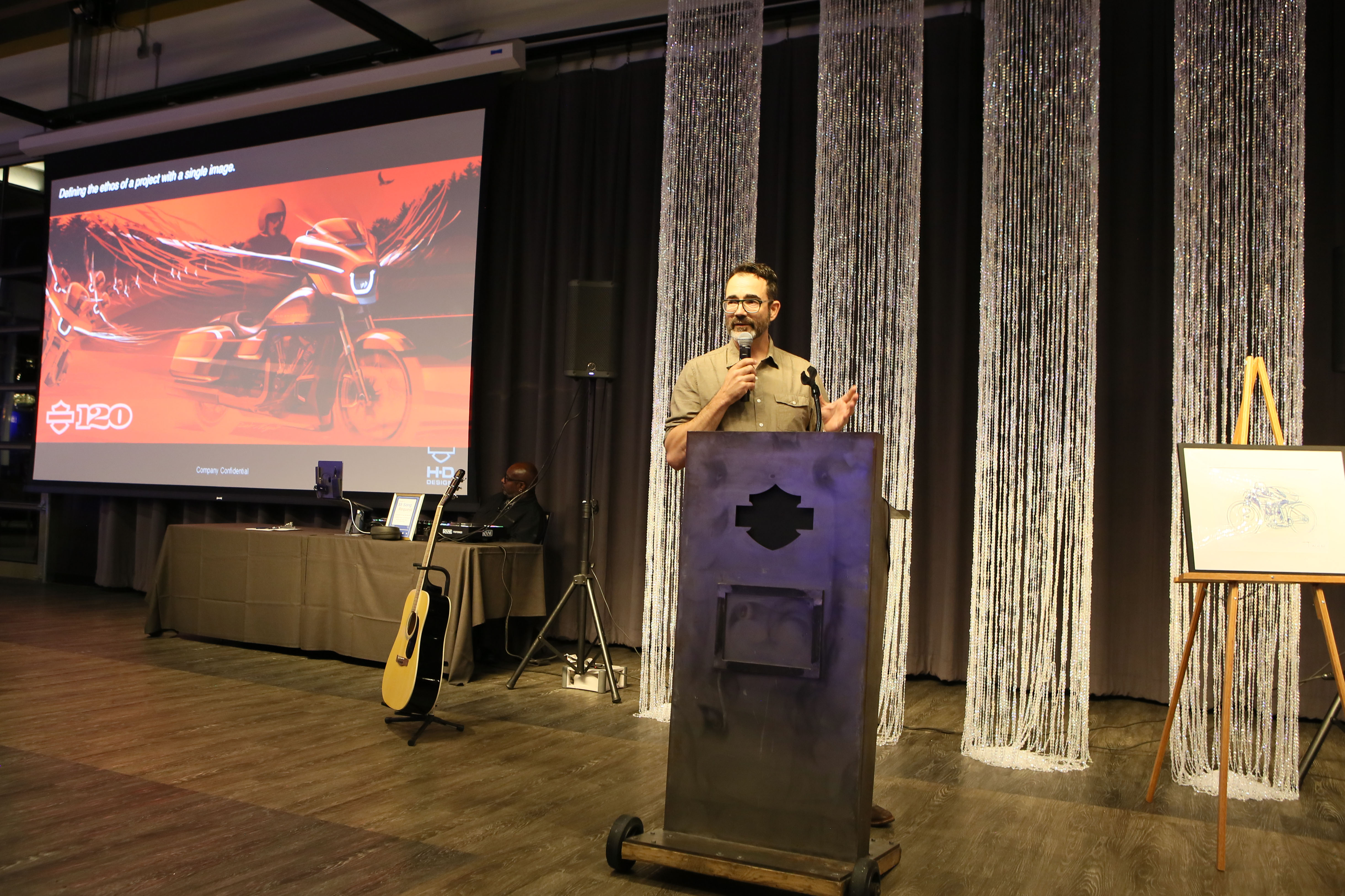 Speaker Bjorn Shuster stands at a Harley-Davidson podium in front of a PowerPoint presentation with an illustration of a motorcycle