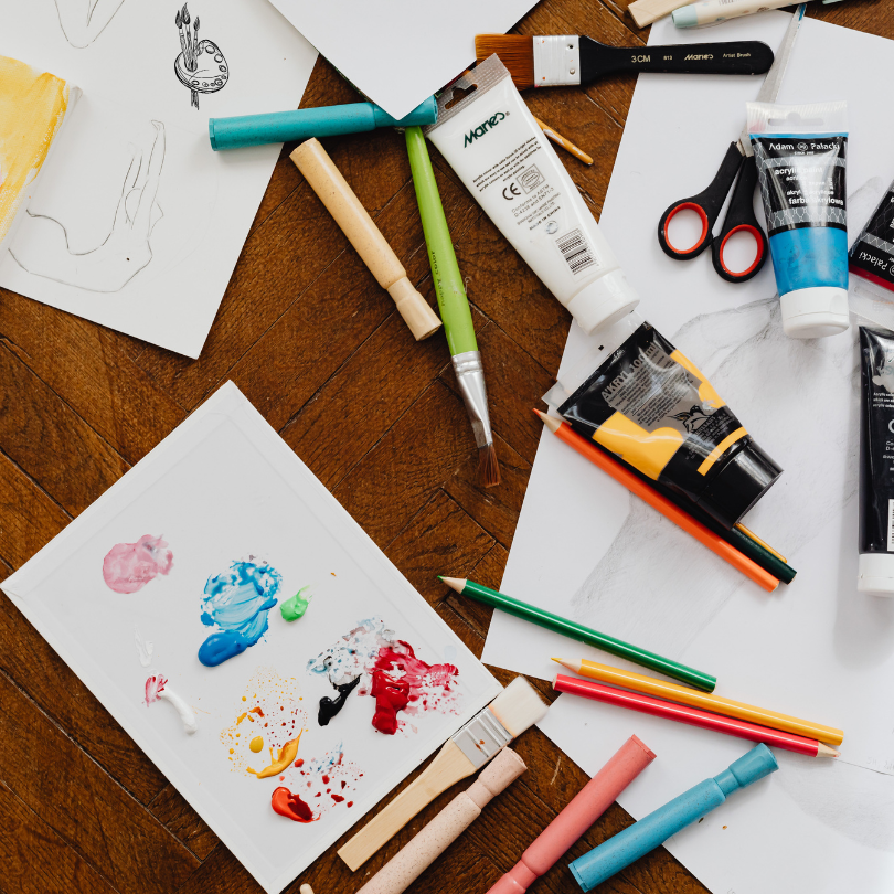 Art supplies spread across a wooden table, including paint tubes, brushes, colored pencils, scissors