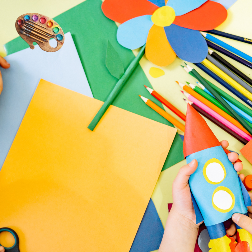 A colorful arts and crafts setup with sheets of paper, colored pencils, scissors, and a paint palett