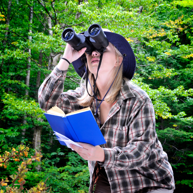 Person outdoors in a wooded area using binoculars while holding an open blue notebook. They are wear