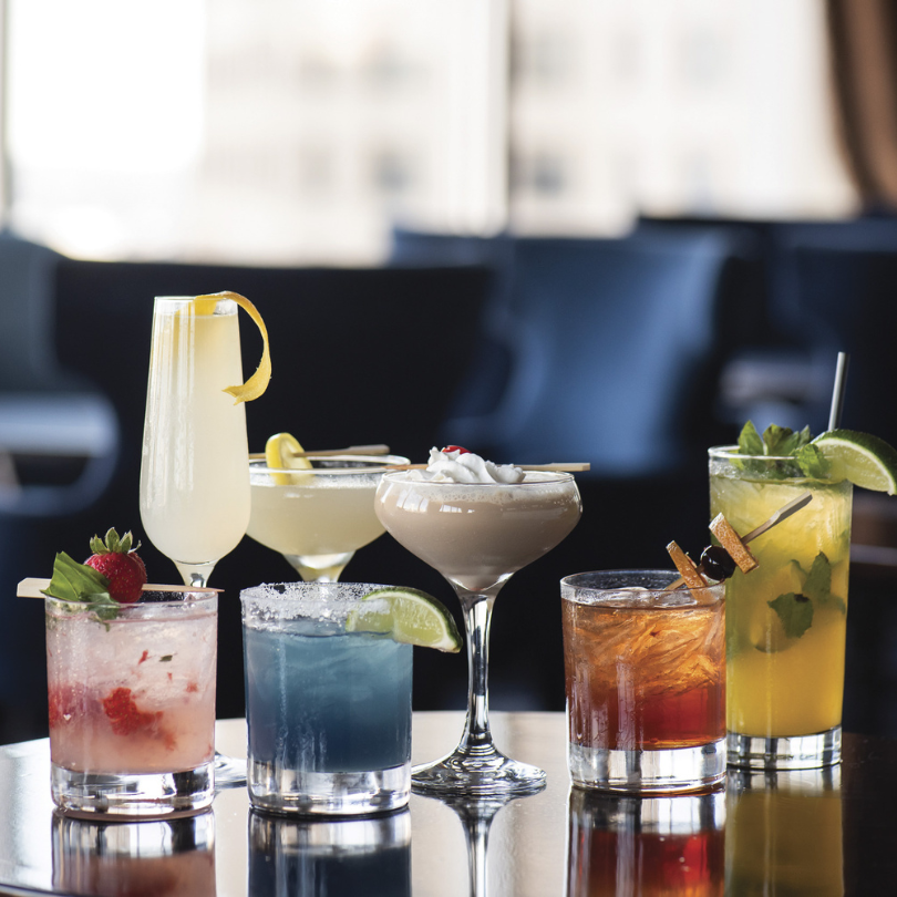 A group of colorful cocktails arranged on a reflective table inside a dimly lit lounge.