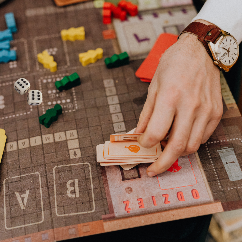 Close-up of a person’s hand placing cards on a tabletop board game. The game board features a grid l