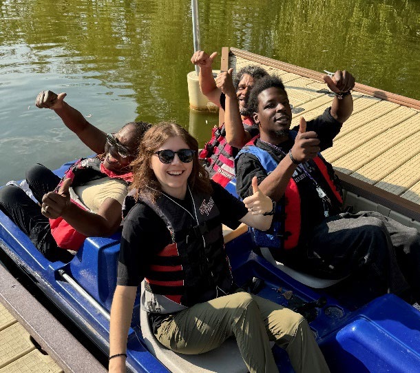 Four people wearing life jackets sit together in a blue paddle boat at the edge of a wooden dock on 