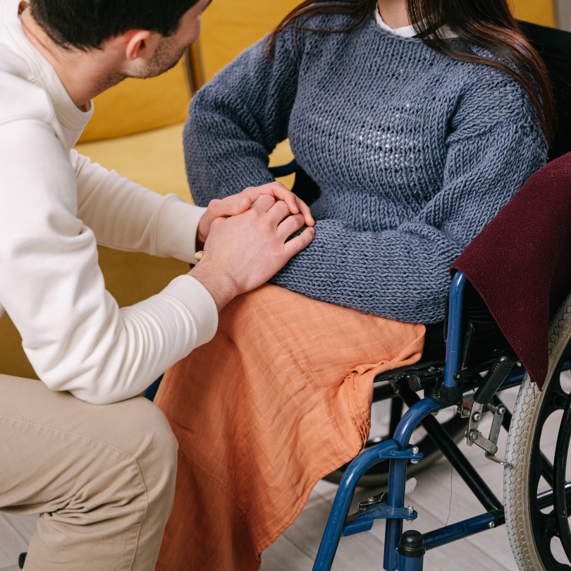 A man kneels beside a woman seated in a wheelchair, gently holding her hands in a supportive gesture