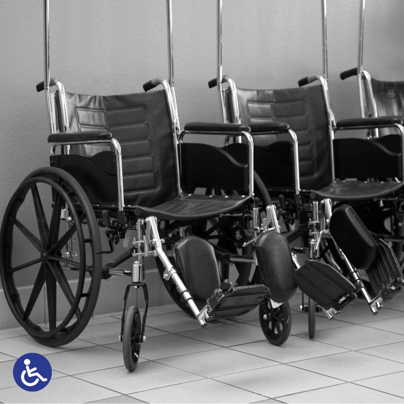 Three manual wheelchairs are lined up indoors against a wall. The image is in black and white, with 