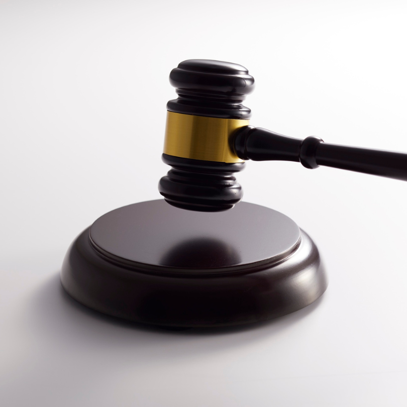 A dark wooden judge’s gavel with a gold band poised above a matching sound block against a plain whi