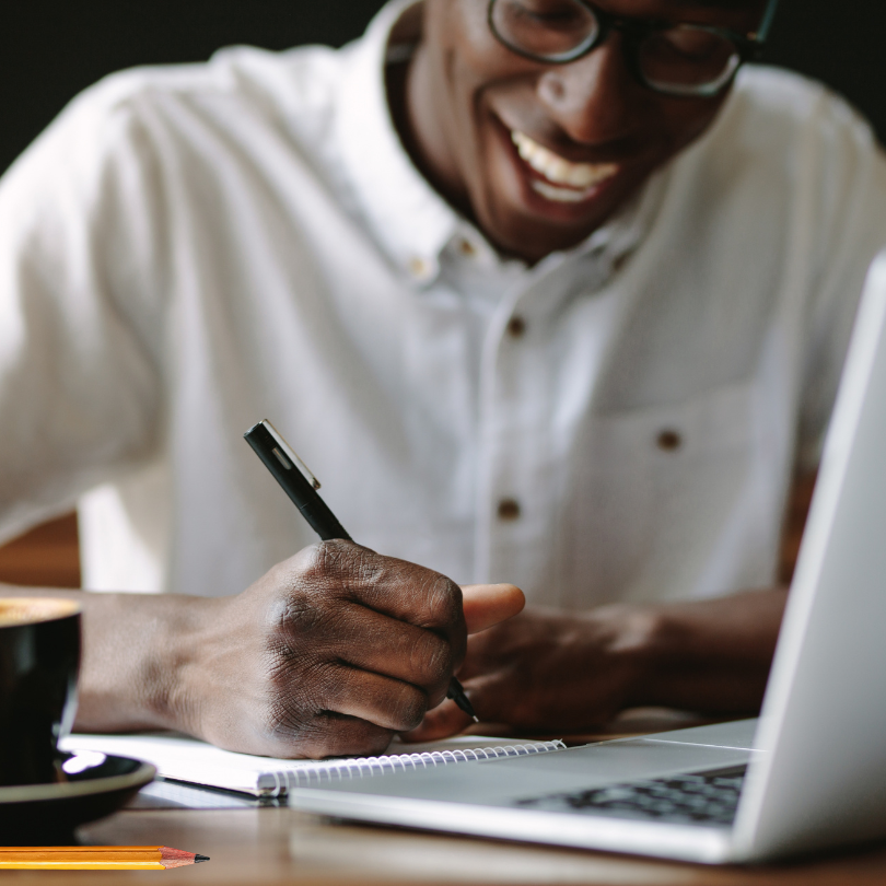 A smiling person writes in a notebook while sitting at a table with a laptop, suggesting a creative 