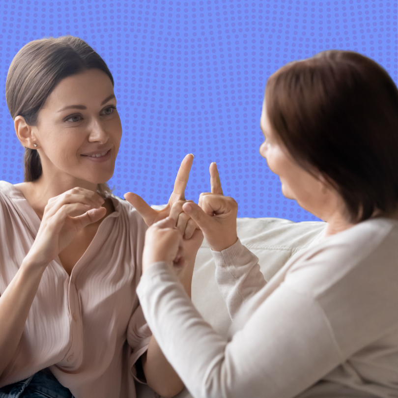 Two women sit facing each other on a couch, smiling and communicating using sign language with their
