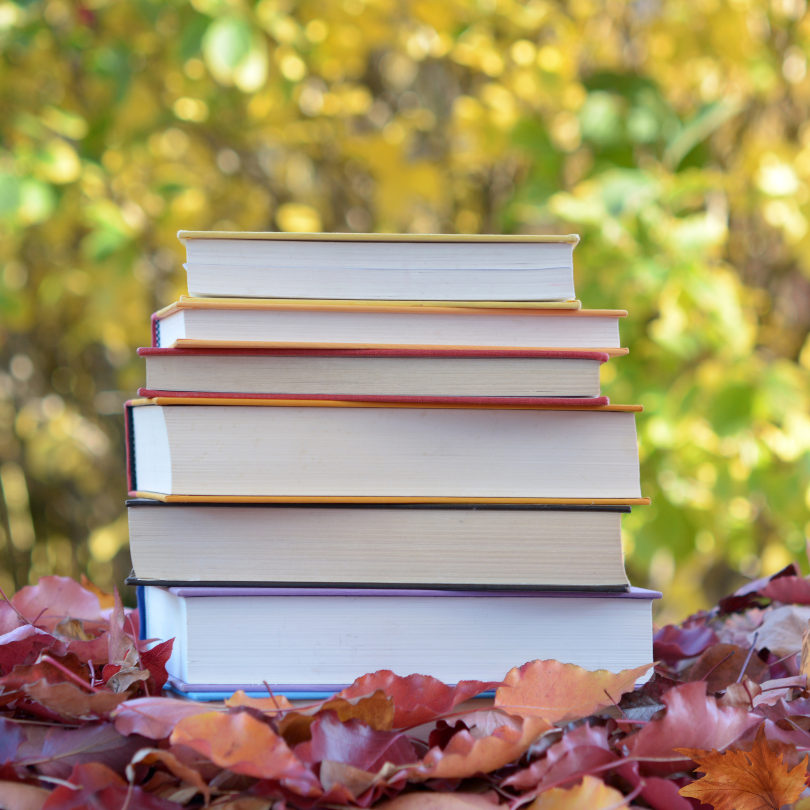 A stack of books sits among colorful red and orange fallen leaves, with softly blurred yellow and gr