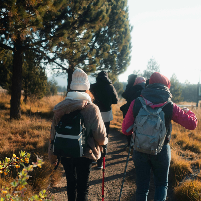 Group of people hiking along a narrow dirt trail through a grassy natural area, seen from behind. Tw