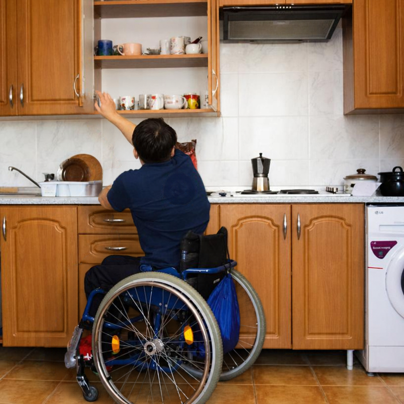 A wheelchair user reaches into an upper kitchen cabinet while seated at a countertop, surrounded by 