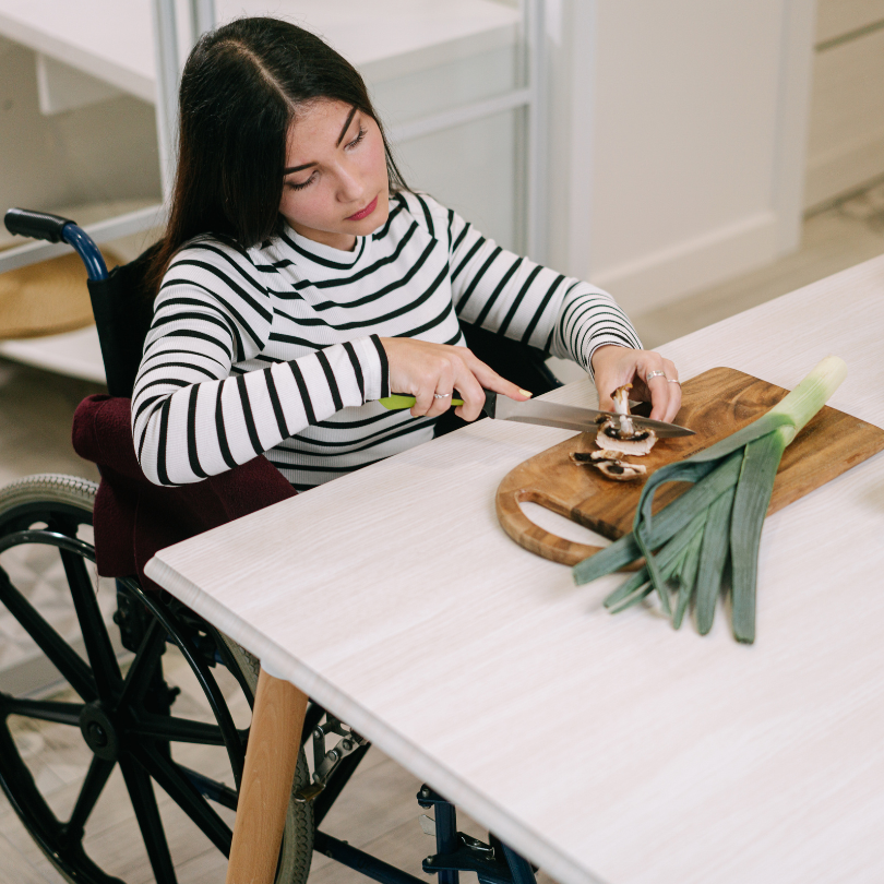 A woman using a wheelchair slices mushrooms on a cutting board at a kitchen table, with a leek besid