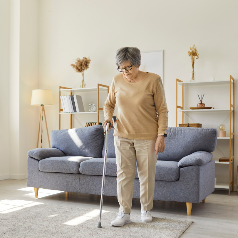 A woman stands in her living room using a cane for support. Behind her is a couch and shelving with 