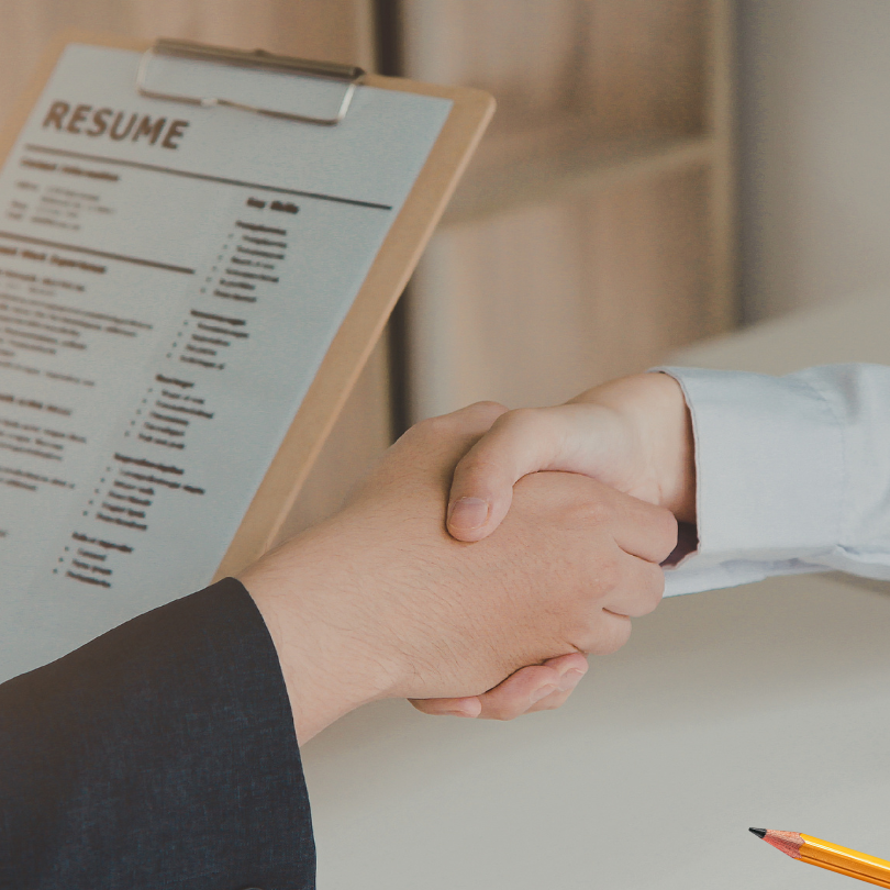 Two people shake hands in a professional setting, with a clipboard holding a resume visible in the b