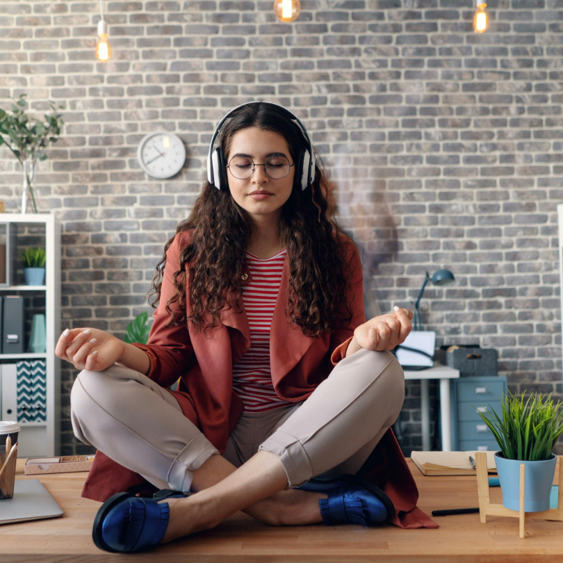 A person with long curly hair sits cross-legged on a desk, eyes closed and wearing over-ear headphon