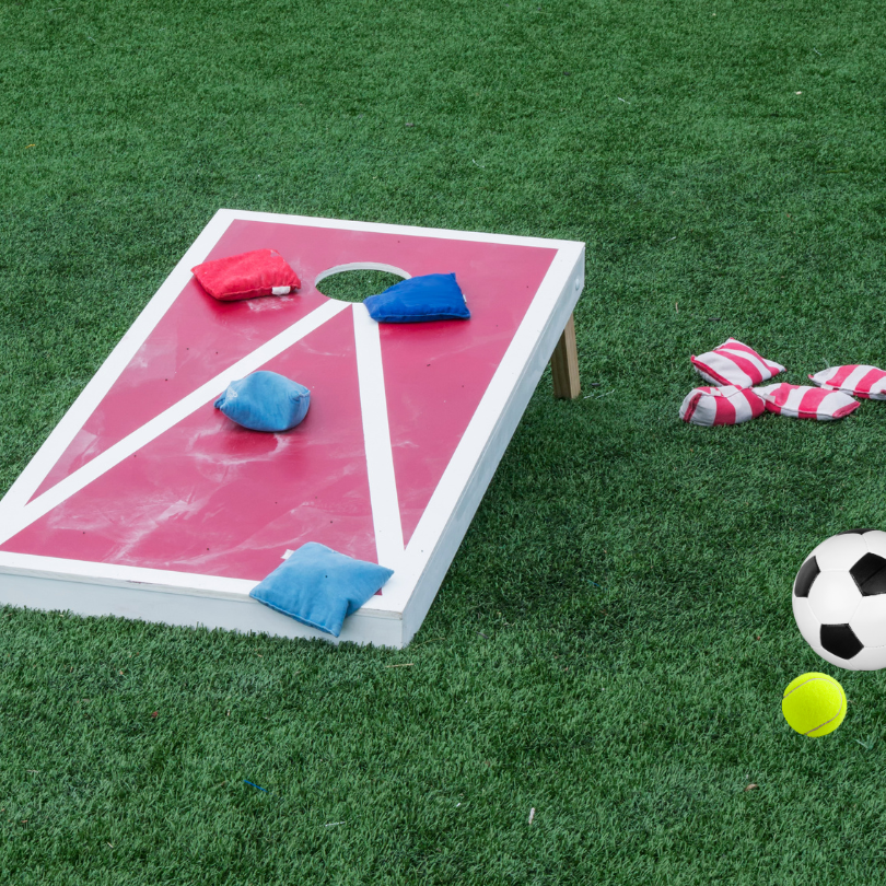 A red and white cornhole board sits on green grass with several blue and red bean bags scattered on 
