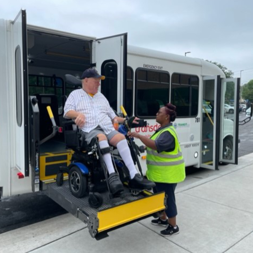 A paratransit bus driver assists a wheelchair user on a lift platform as they board a Transit Plus b