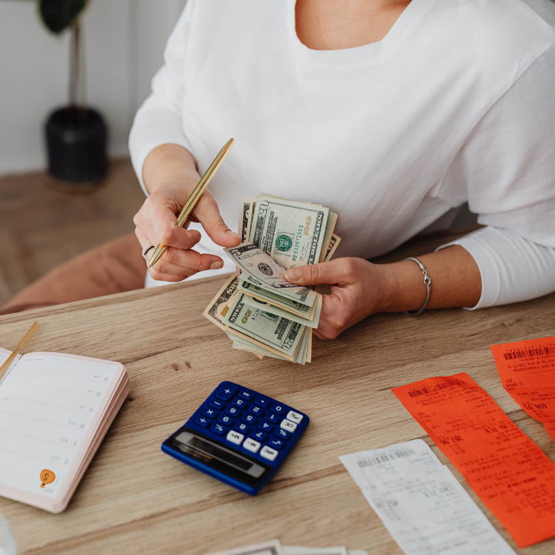 A person sitting at a table counts U.S. dollar bills while holding a pen, with a calculator, receipt