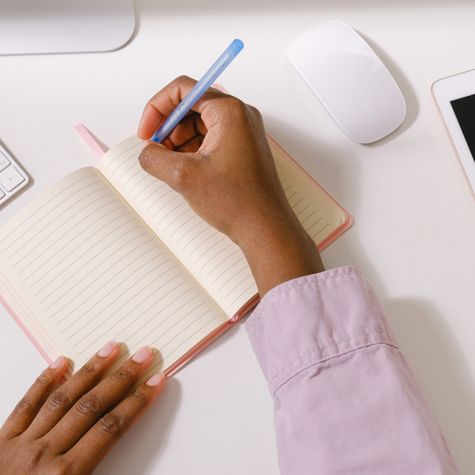 Two hands on a desk with one hand writing in a notebook