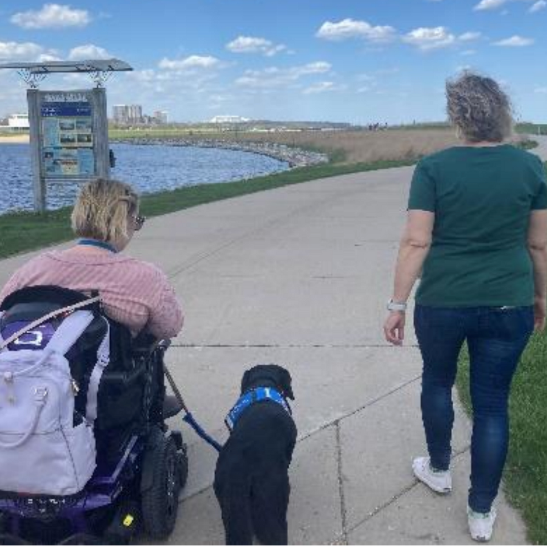 A woman using a wheelchair and service dog walks with another woman on a nature path along a lake