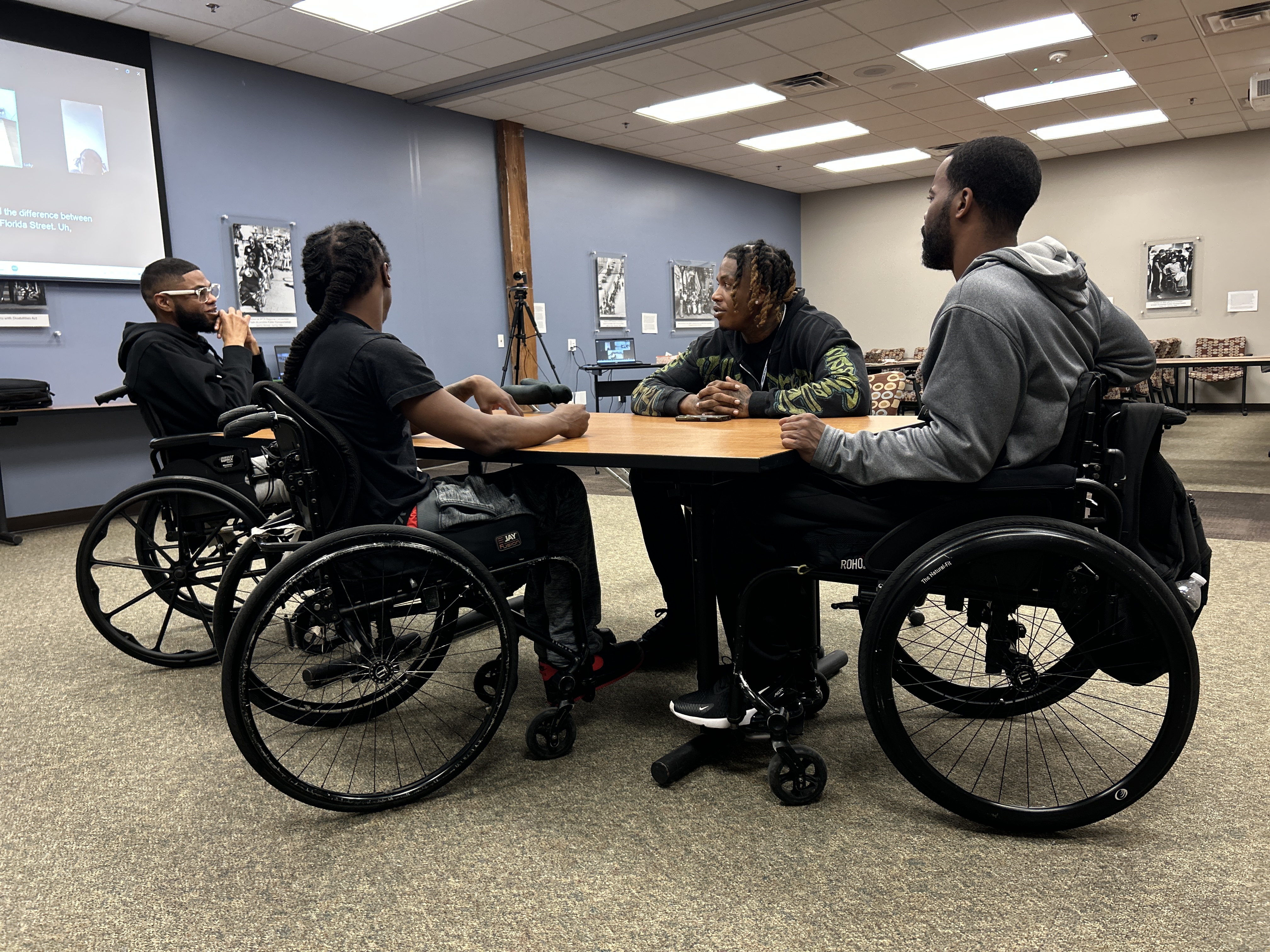 A group of young men sit around a table. Three are in wheelchairs and one is seated in a traditional chair.
