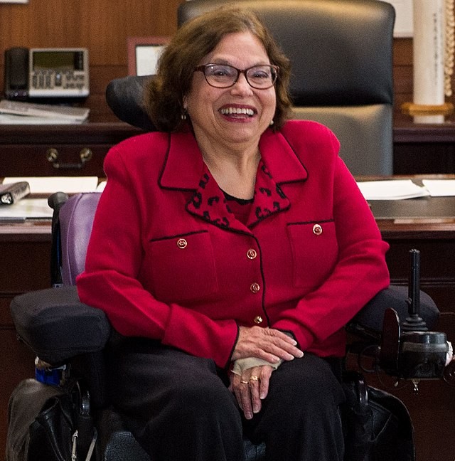 A woman in a purple suitcoat seated in a power wheelchair smiling.