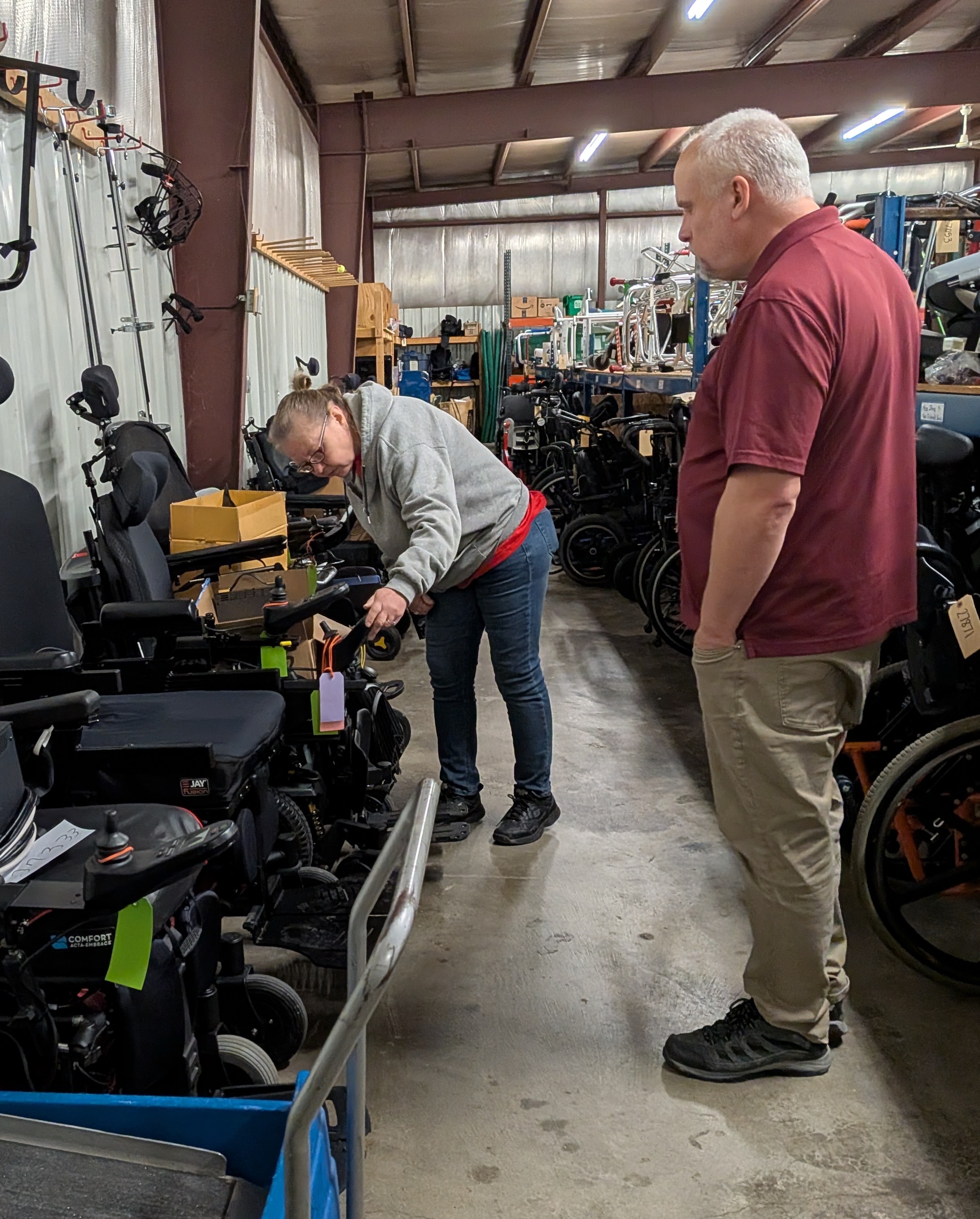Two adults stand inside a large warehouse filled with rows of power wheelchairs and mobility equipment. One person leans over a power chair, examining its controls, while the other watches. The space has metal beams, industrial lighting, and shelves stocked with additional assistive devices and parts in the background.