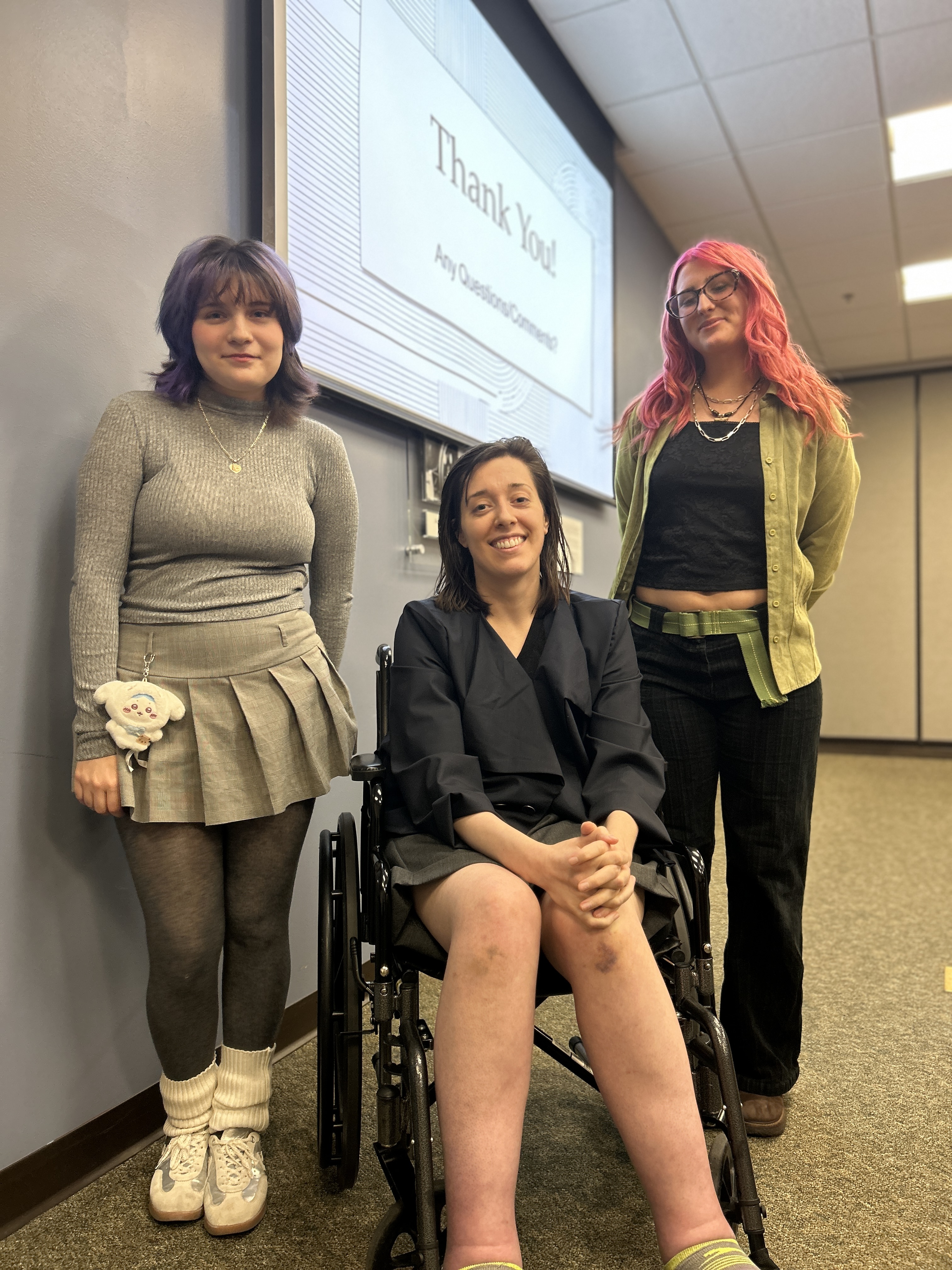 ​Three young women pose for a photo. A student on the left stands with a grey sweatshirt. A young woman is seated wearing a dark coat. A student is standing with pink colored hair.