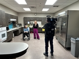 A young woman with purple pants addresses a camera. Beside her is a woman using a wheelchair. They are standing in the kitchen at Independence First.