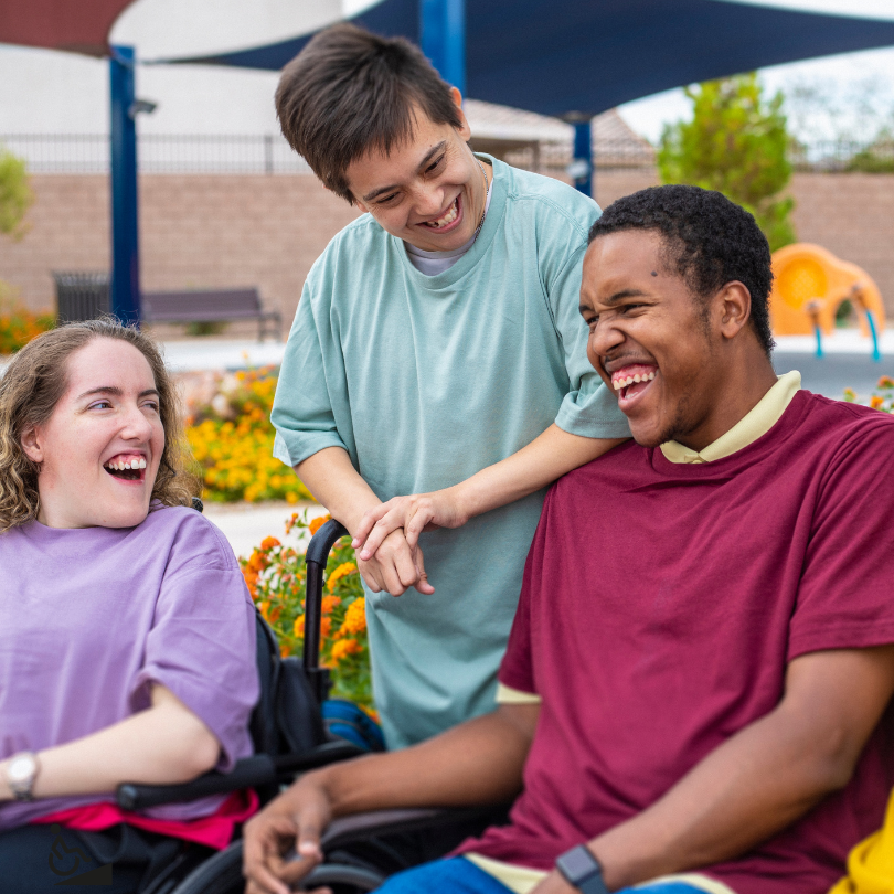 Three young adults with disabilities laugh and talk together outdoors at a park. One person sits in 