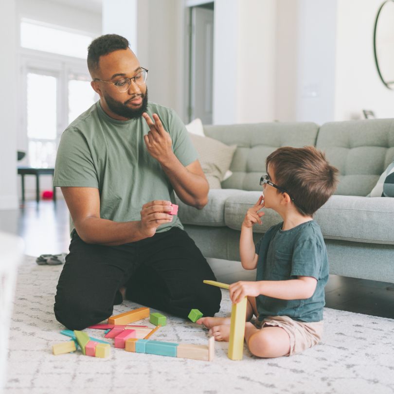 An adult uses ASL to communicate with a child who is playing with blocks on a living room floor