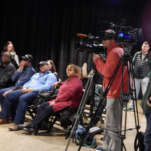 Several people are sitting in chairs with a camera crew nearby.