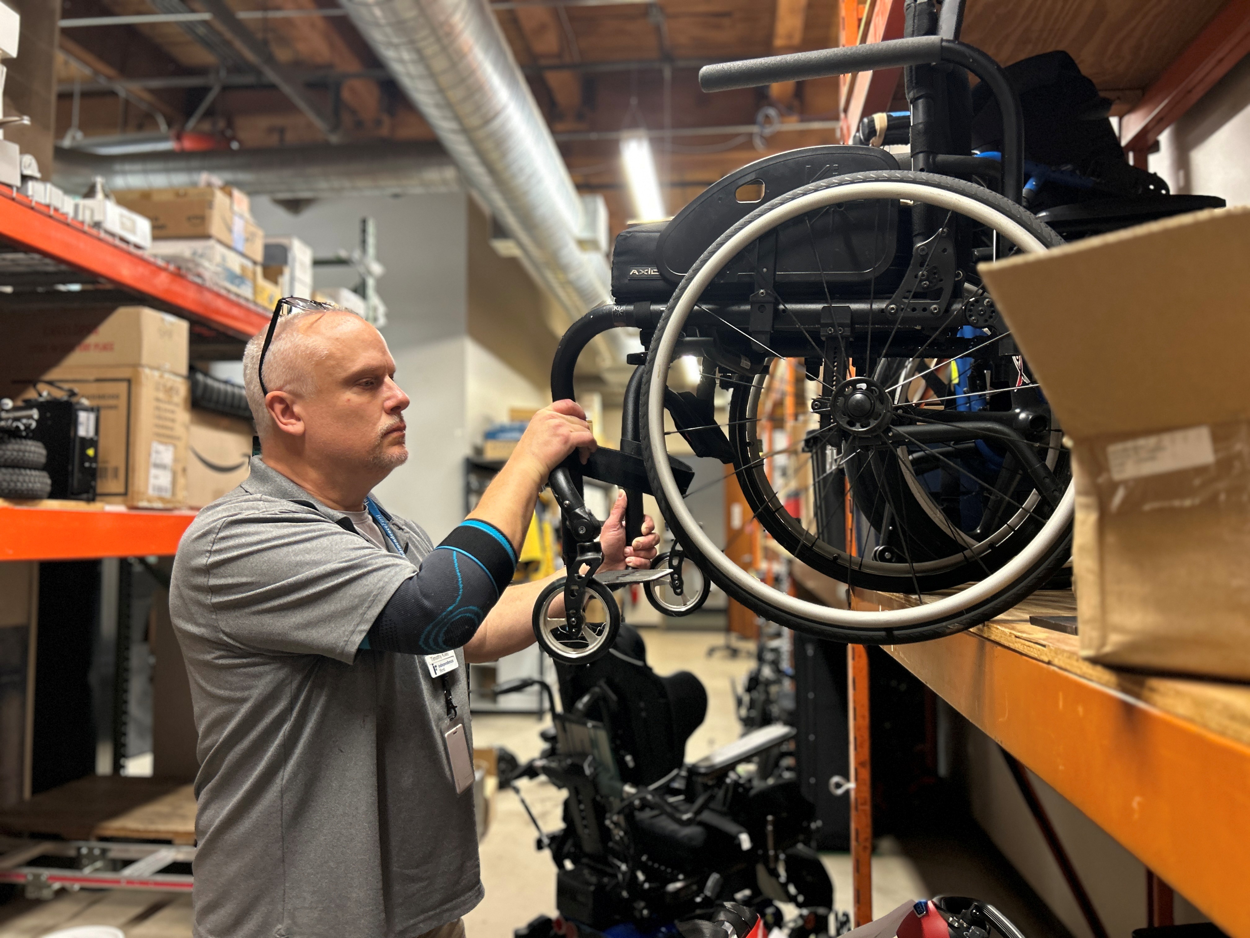 An assistive technology specialist inspects and adjusts a manual wheelchair stored on industrial shelving in a workshop, surrounded by mobility equipment and repair supplies used for refurbishment and maintenance.