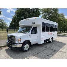 White Milwaukee County Transit Plus paratransit van parked in a paved lot near a grassy area and trees on a sunny day. The accessible shuttle bus has large side windows and the Transit Plus branding on the side.