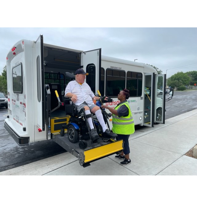 A Milwaukee County Transit Plus paratransit van is parked with its wheelchair lift deployed. A wheelchair user sits on the lift platform while a driver wearing a reflective safety vest assists them as they board the vehicle. The accessible van is parked along a sidewalk on an overcast day.