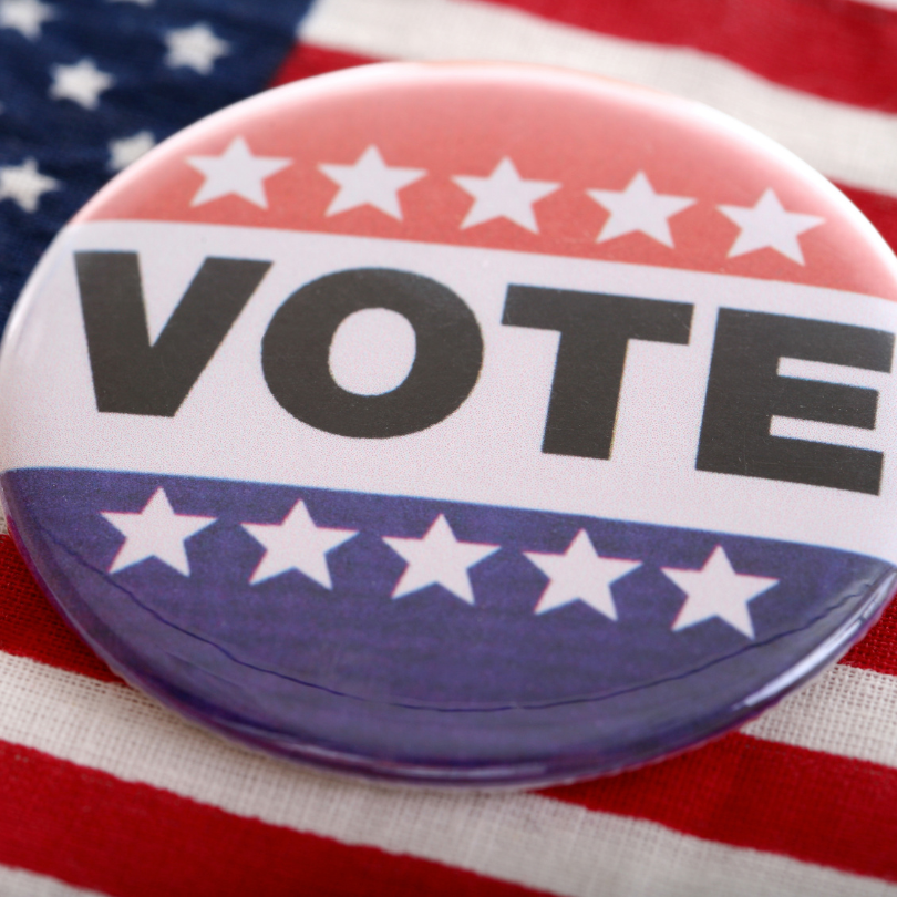 Round red, white, and blue “Vote” button with white stars resting on an American flag background.