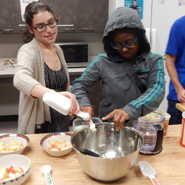 A woman and a student cooking in a kitchen using a bowl and various ingredients.  