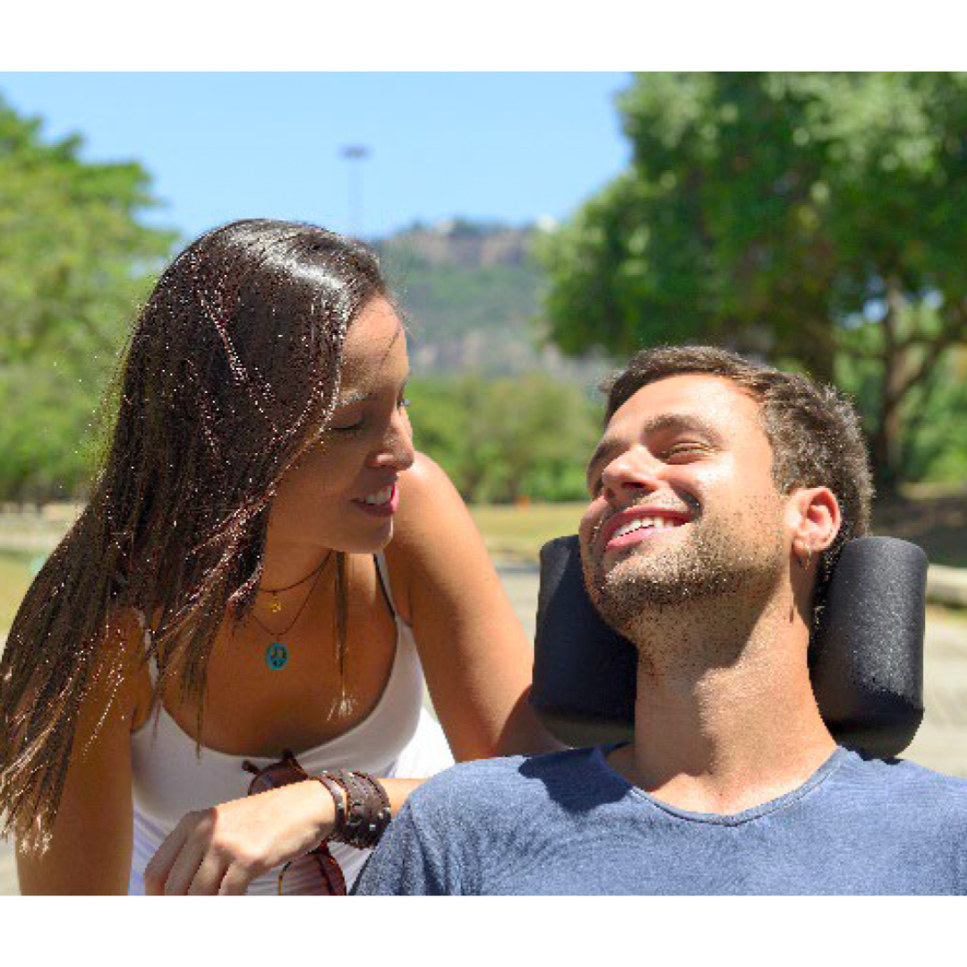 Young man in a power wheelchair smiles in the sun with a young woman leaning on his shoulder